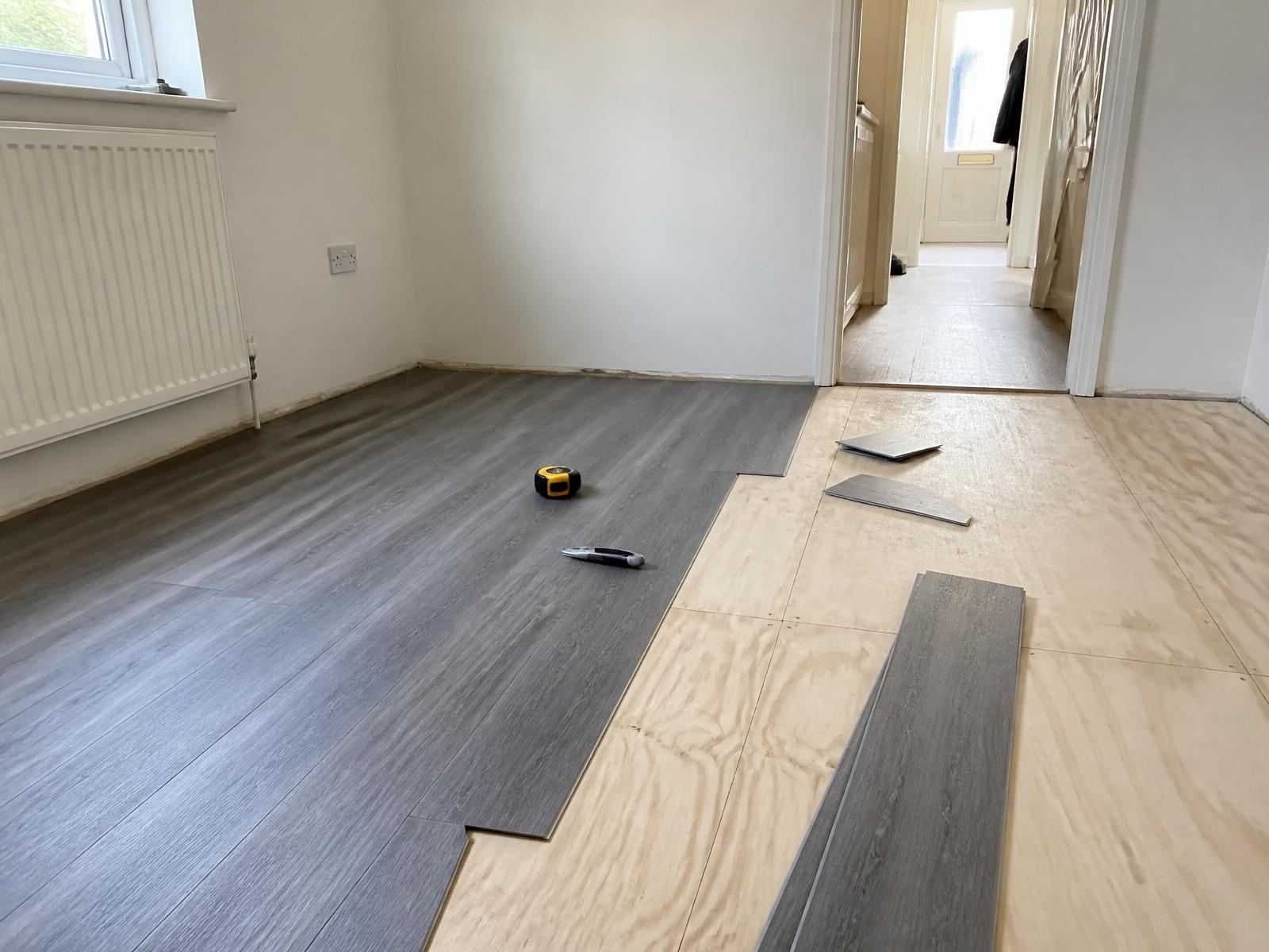 Vinyl plank flooring half laid over plywood with offcuts and tools on the floor